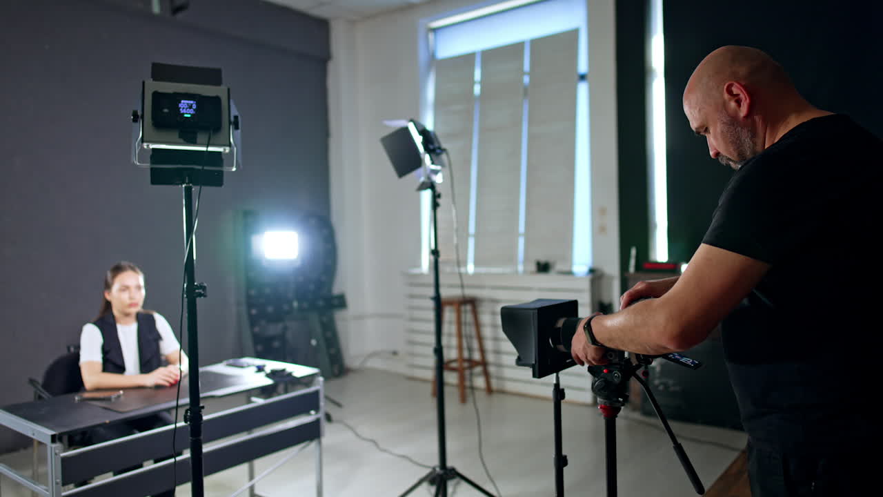 Young woman sits at the table. Cameraman sets the equipment for footage around the lady. Studio shooting.