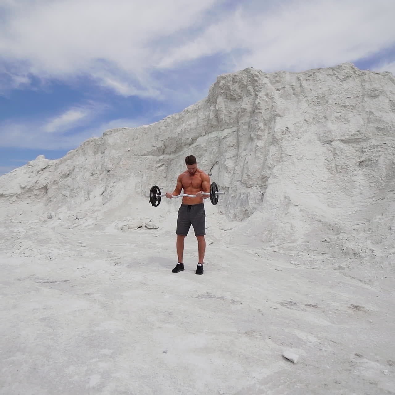 Young athlete lifting heavy barbell on the natural mountain background. Concentrated man engaged with barbell outdoors in a summer day.