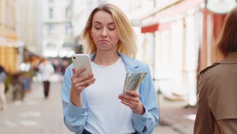 Mujer-Rubia-Feliz-Contando-Dinero-En-Efectivo-En-Dólares,-Usa-La-Aplicación-De-Calculadora-De-Teléfono-Inteligente-En-La-Calle-De-La-Ciudad