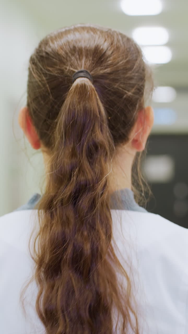 vista posterior del pasillo de una clínica, primer plano de una técnico con coleta y bata blanca, paseo lento por un pasillo esterilizado, pelo ondulado recogido, iluminación cenital suave, puertas y señalización desenfocadas