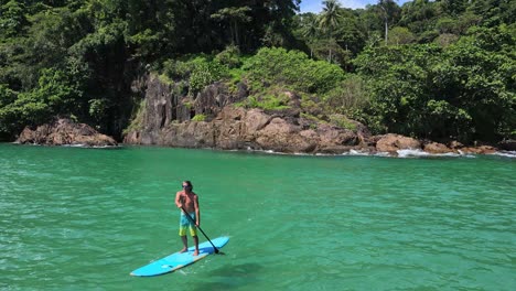 backwards dolly aerial drone shot of caucasian man exercising on a sup paddle board in turquoise tropical clear waters, with tropical jungle palm trees and coastline in thailand