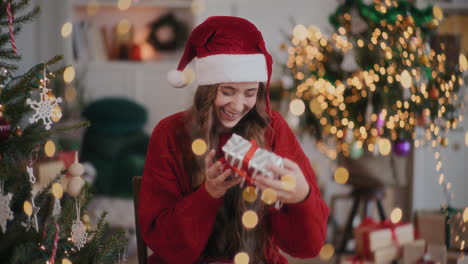 mujer emocionada en sombrero de santa jugando con caja de regalos en casa decorada