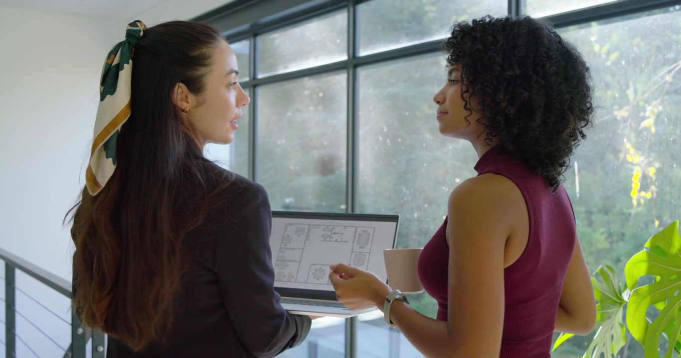 Diverse female coworkers tilting laptop to show plan, pointing at window and evaluating light