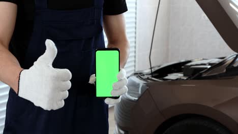 un mecánico de automóviles con un mono azul sostiene un teléfono con una pantalla verde en su mano y muestra un gesto de aprobación. servicio de reparación de automóviles en el garaje.