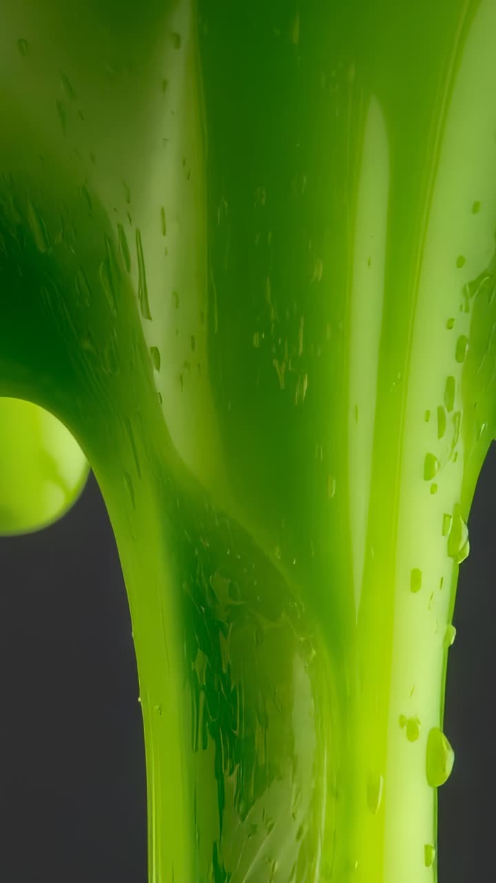 Vertical video: Entering green liquid ribbon flowing down against gray backdrop, splitting droplets