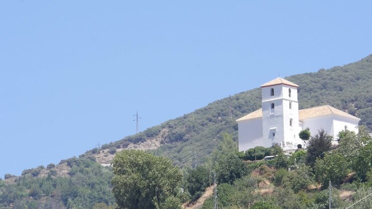 Vista di una chiesa rurale andalusa, con una facciata bianca e un campanile sormontato da tegole arabe. La chiesa si erge su una collina verde nella regione dell'Alpujarra di Granada.