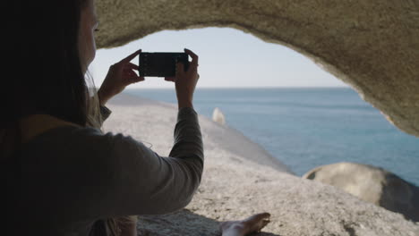 un primer plano de una joven turista tomando fotos usando tecnología móvil de teléfono inteligente disfrutando de la hermosa vista del océano sentada en una cueva junto al mar relajándose