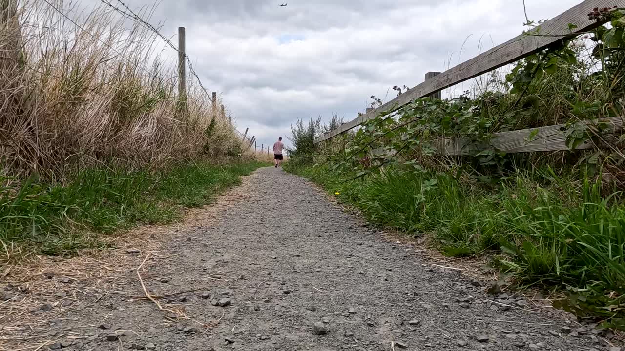 Low-angle camera moves steadily along a gravel path bordered by grass and wooden fencing, under overcast skies, creating a tranquil, natural atmosphere