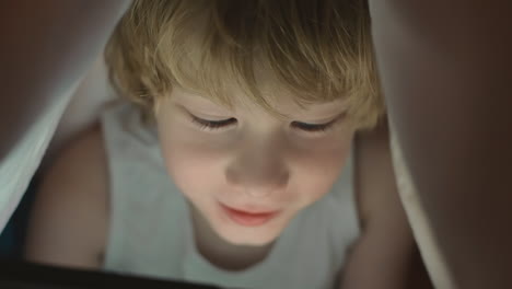 little boy using a tablet covered by the bed sheet at night