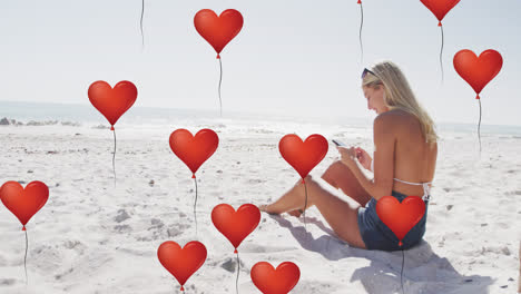 animation of red heart love balloons digital icons over woman using smartphone on beach