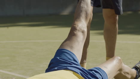 vertical shot of a senior sportsman lying down on pitch while his friend stretching his leg