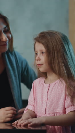 cheerful woman puts pill to adorable little daughter mouth to cure pain and hugs girl at small table in semi-dark room at home close view slow motion