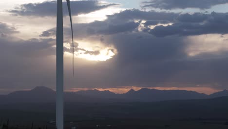 cordillera en la distancia con sol poniéndose detrás de nubes de tormenta con aspas de turbina eólica de giro lento en primer plano