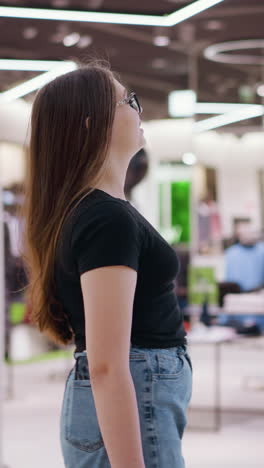배경에 보케 빛이 있는 쇼핑몰을 고 있는 안경 입은 여자의 측면 전망, 의류 상점과 현대적인 인테리어 디자인, 트렌디한 패션, 그리고 스타일리시한 쇼핑 경험