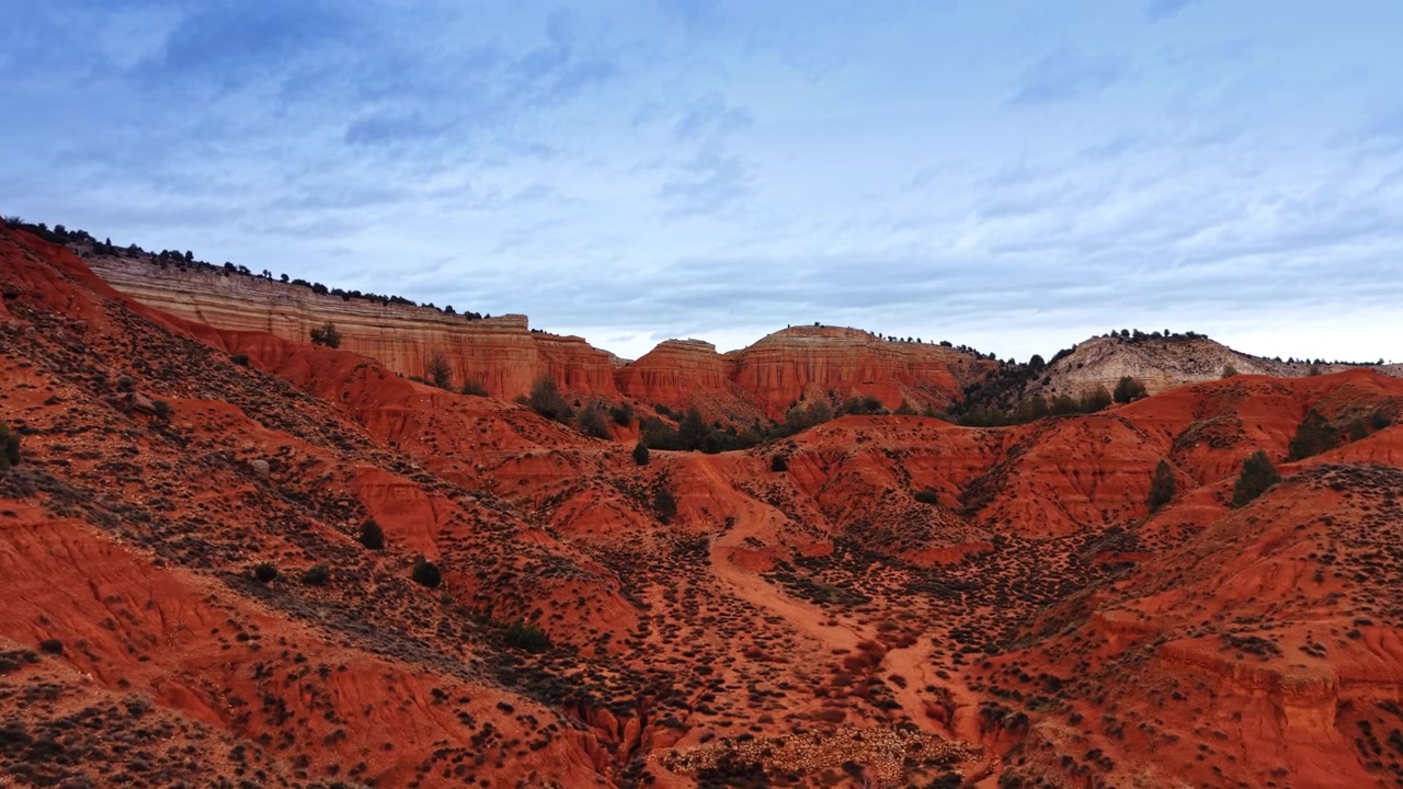 Badlands de roca roja con acantilados estratificados bajo cielo nublado, España. Vista a nivel del suelo de mesetas erosionadas, arbustos dispersos y sendero sinuoso.