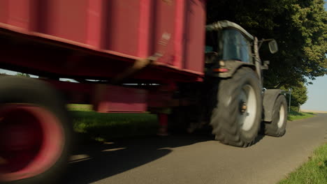 a red agricultural tractor with a large trailer speeds along a sunny countryside road, passing under the shade of a tree in a rural setting