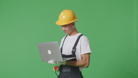 side view asian man painter wearing safety helmet using a laptop while standing in the green screen background studio
