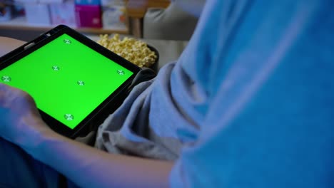 de cerca, un hombre sosteniendo una tableta con pantalla táctil verde y comiendo palomitas de maíz en el sofá en casa.
