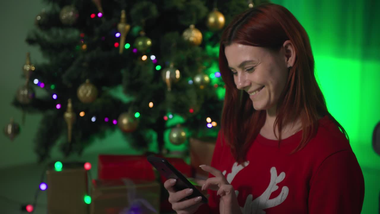 새해 전날 휴대전화를 가진 행복한 소녀, 젊은 여성은 카메라에 녹색 화면과 선물을 가진 스마트폰을 보여줍니다. 밤에는 많은 불빛이있는 크리스마스 트리의 배경에