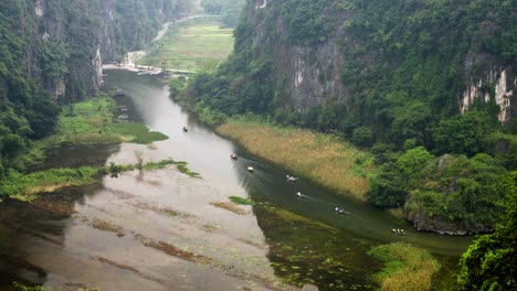 from mua cave's elevated viewpoint, small boats navigate through a river flanked by towering limestone cliffs and lush vegetation in vietnam's trang an grottoes, creating a mystical landscape