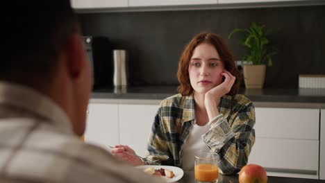 Over-the-shoulder-a-young-adult-girl-with-brown-hair-and-a-bob-hairstyle-communicates-with-her-boyfriend-with-Black-skin-color-during-their-breakfast-together-in-a-modern-kitchen-in-an-apartment