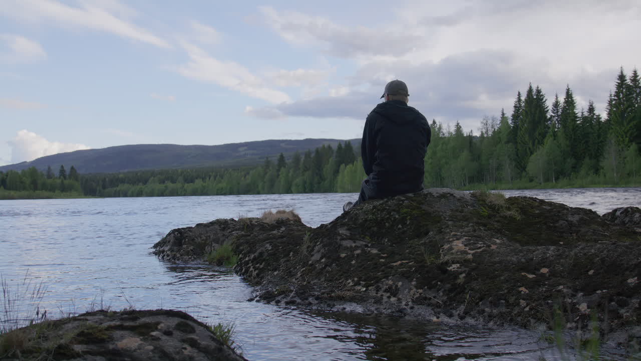 Person sitting alone on mossy rock island by wide flowing river with hand waving in peaceful gesture, surrounded by forest and mountains under blue sky in remote wilderness setting