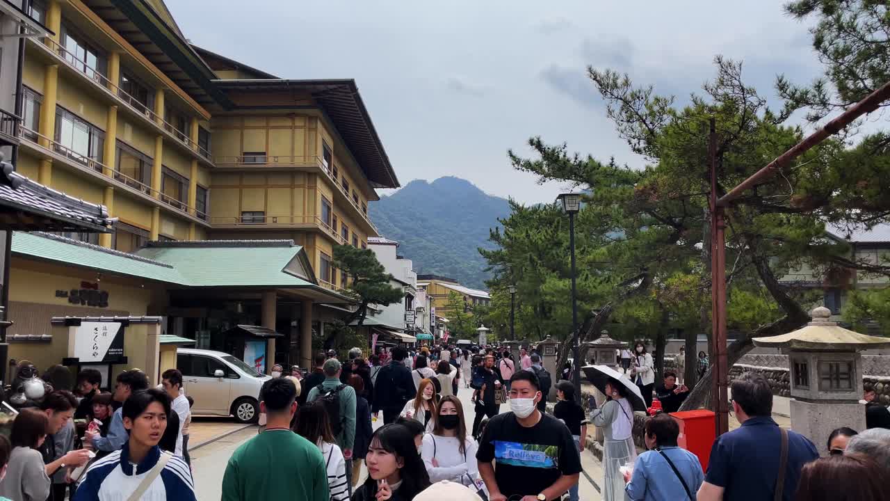 Crowds of tourists walk through Miyajima Omotesando Street between apartments and trees on an overcast day in Japan, with Mount Misen in the distance