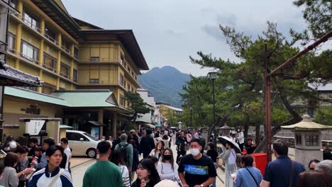 crowds of tourists walk through miyajima omotesando street between apartments and trees on an overcast day in japan, with mount misen in the distance