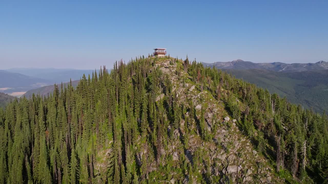 Un drone aereo che sorvola una torre di avvistamento incendi isolata nell'Idaho settentrionale, con persone che si godono il paesaggio montano remoto nel tardo pomeriggio e una città in lontananza