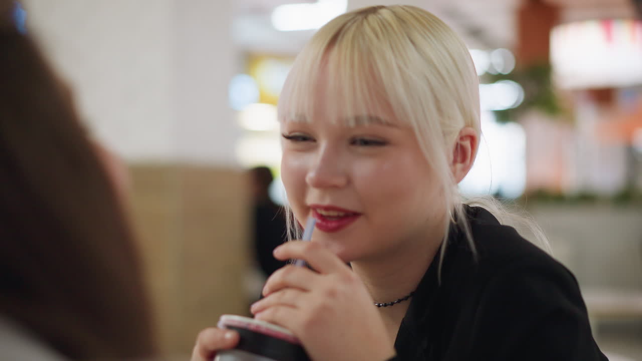 Smiling young lady with blonde hair interacts warmly with friend during relaxed cafe conversation, holding straw while laughing, expressing joy, connection, and lighthearted mood