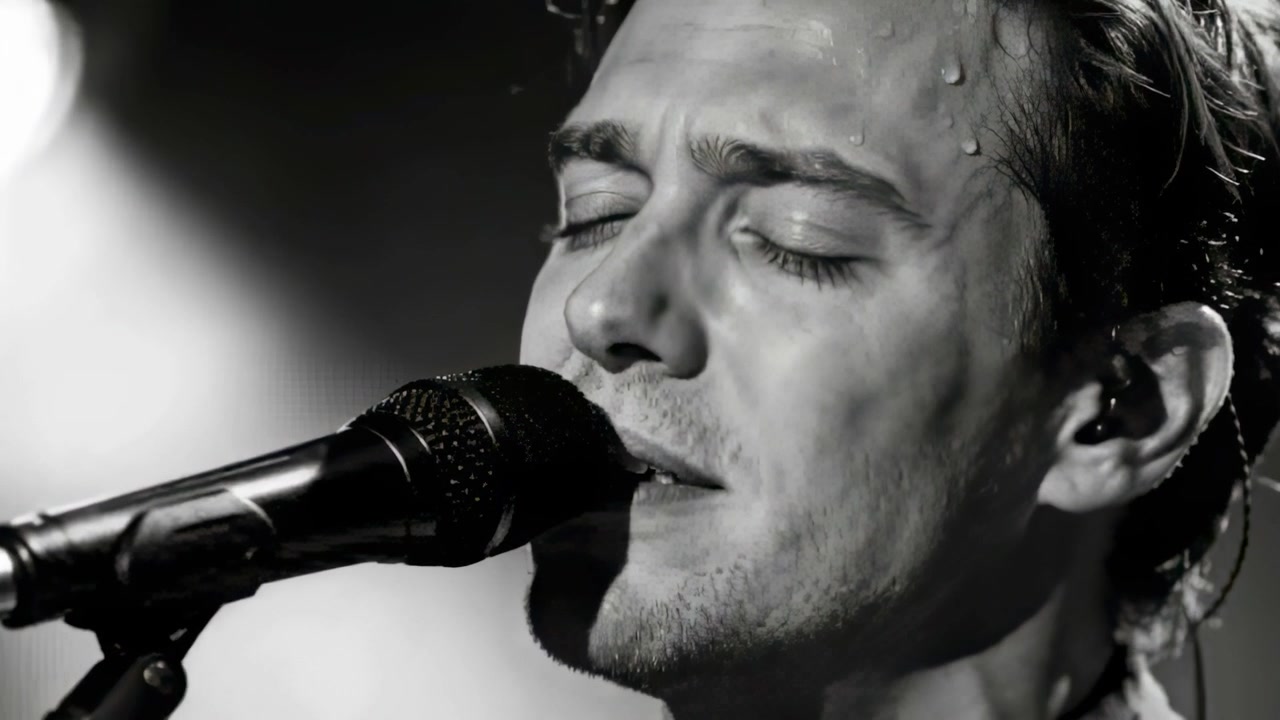 Singing man onstage after cue, leaning into mic, showing collared shirt, in-ear monitor and sweat