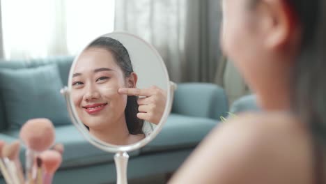 거울에 반사 된 아름다운 젊은 아시아 여성이 얼굴 크림을 적용하고 집에서 메이크업을 적용하는 동안 미소 짓고 있습니다.