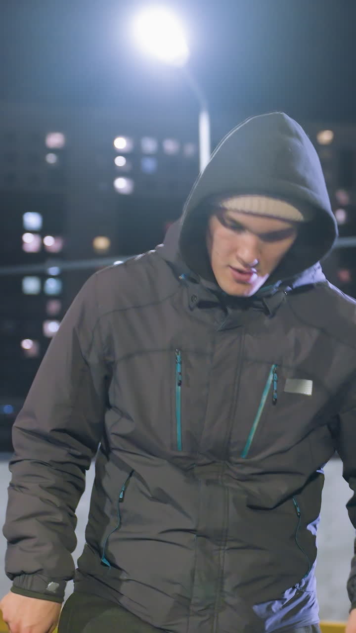 Footballer in hoodie joggling football during nighttime practice on urban sports field surrounded by illuminated high-rise buildings and bright streetlights