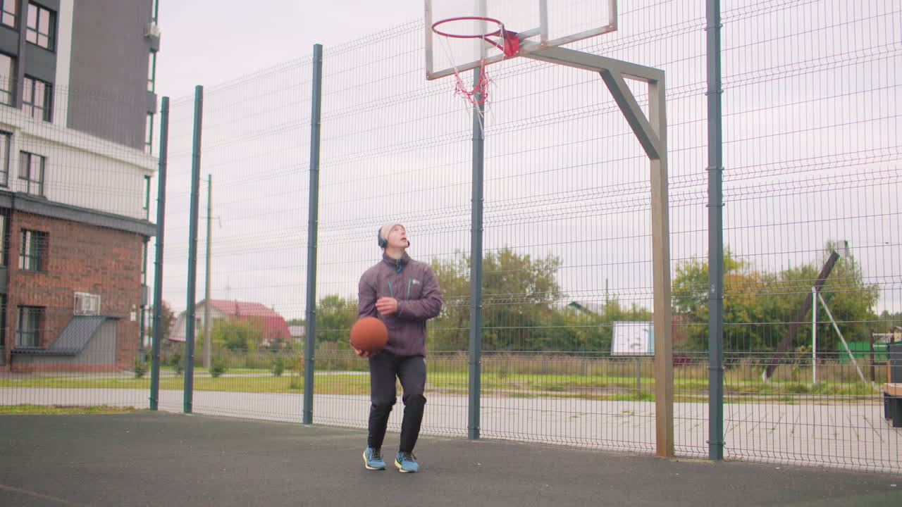 Uomo anziano bianco che cammina con un pallone da basket in un campo urbano recintato, esercizi di riscaldamento leggeri e palleggio leggero, indossa giacca e scarpe da ginnastica, ritmo rilassato, edifici del quartiere sullo sfondo, cielo nuvoloso