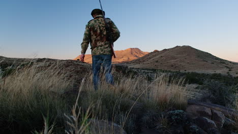 hunter with hunting rifle over shoulder walks to lookout point, vista over karoo landscape at sunset