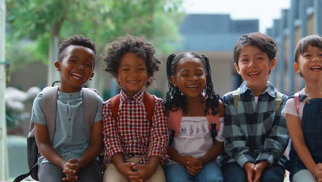 retrato de alumnos de escuelas primarias multiculturales sonrientes sentados en la pared al aire libre en la escuela
