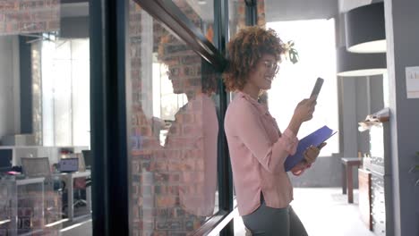 happy biracial casual businesswoman using smartphone standing in office corridor, slow motion