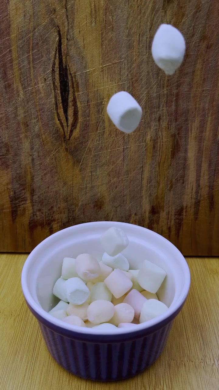 Seramik bir kaseye düşen, kabı yavaşça dolduran ve rustik ahşap bir arka plana karşı eğlenceli yiyecek hareketini yakalayan marshmallowların yakın çekimi