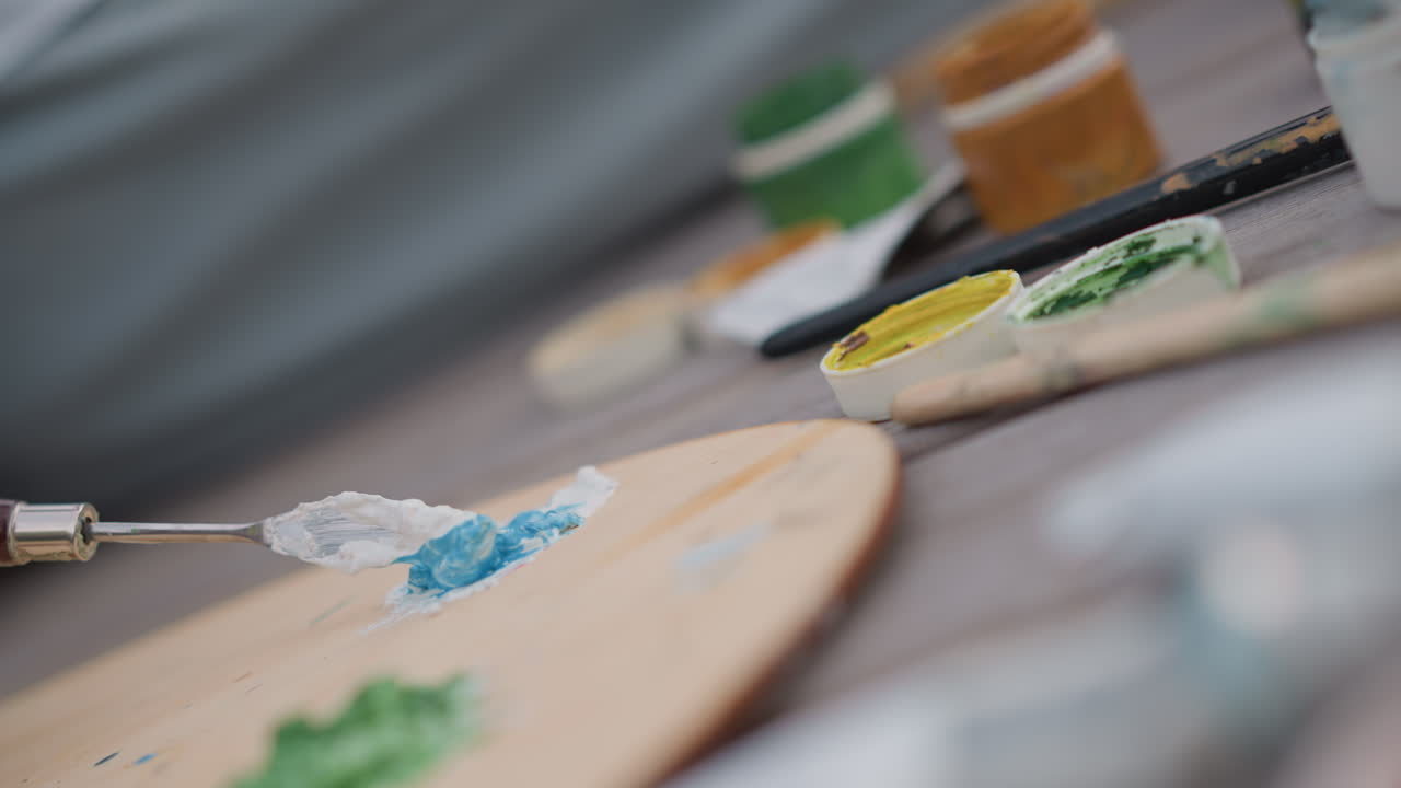 primo piano di un coltello che spalma spessa vernice blu su una palette di legno, pittore in studio che lavora con una spatola, texture a impasto lucido, barattoli di colore sullo sfondo, pennellate dinamiche, movimento tattile, messa a fuoco