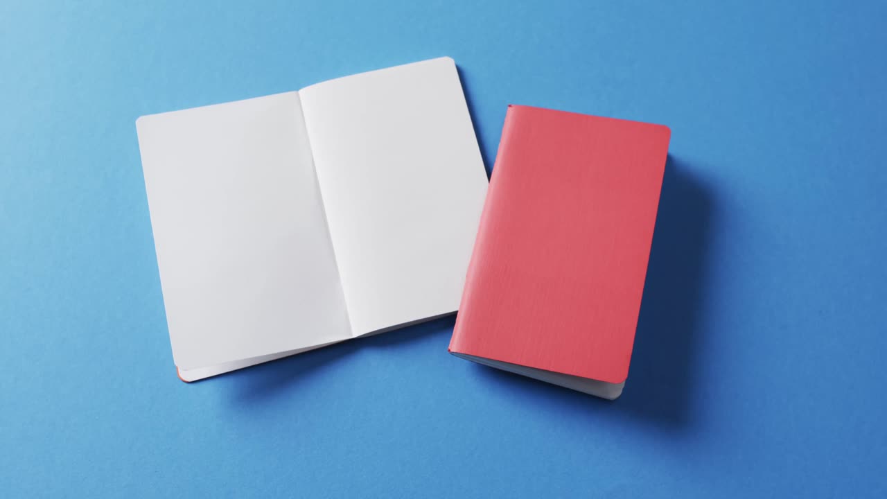 Close up of open blank book and red notebook with copy space on blue background in slow motion