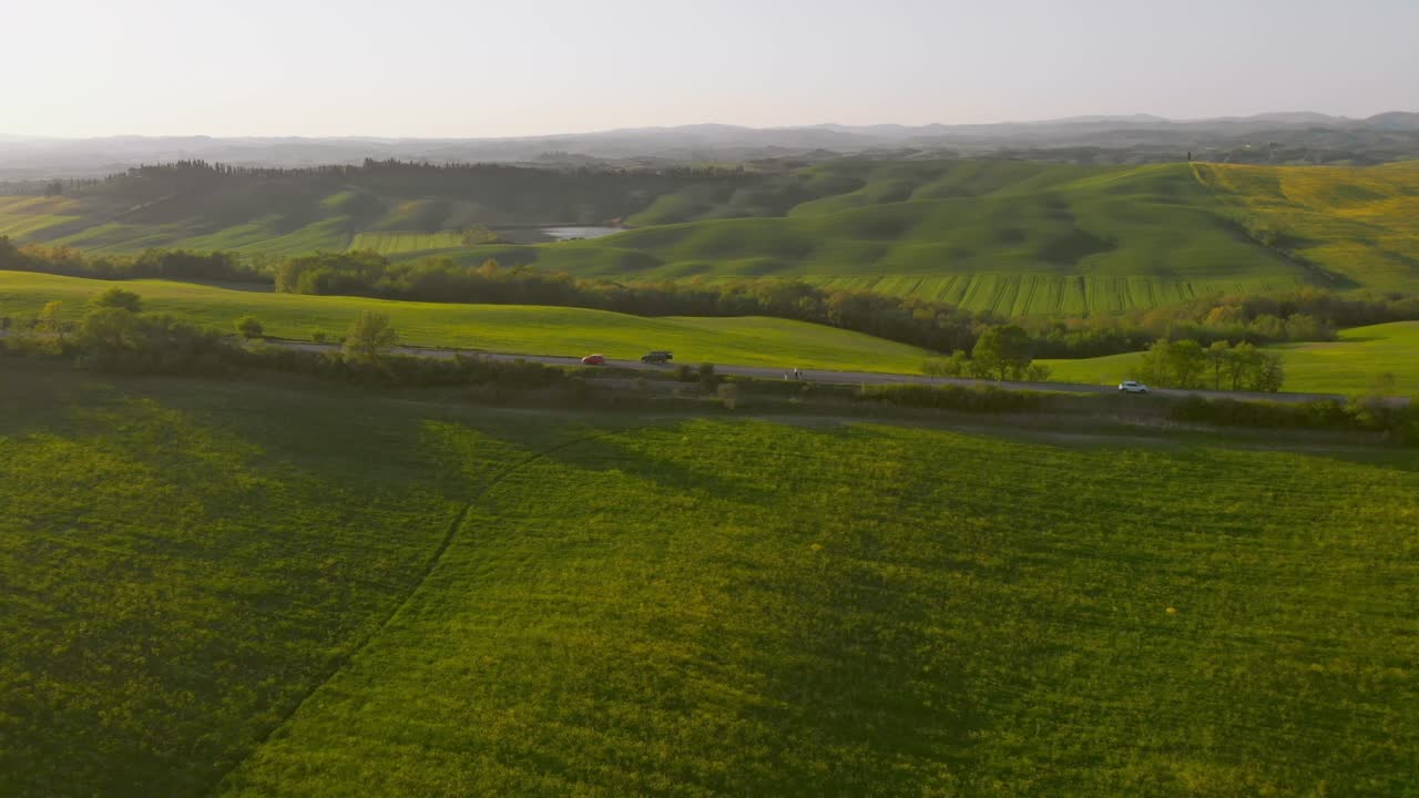 Aerial wide shot of lush green fields and rolling Tuscan hills with winding road and distant layered mountains