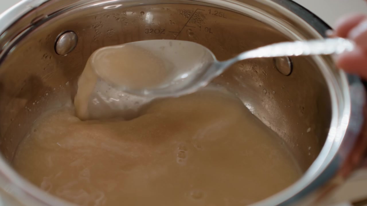 Top-down close-up of stainless steel saucepan with smooth light-brown sauce being stirred and scooped by spoon, forming concentric ripples, glossy trails, and thickening surface, with sauce, stirring