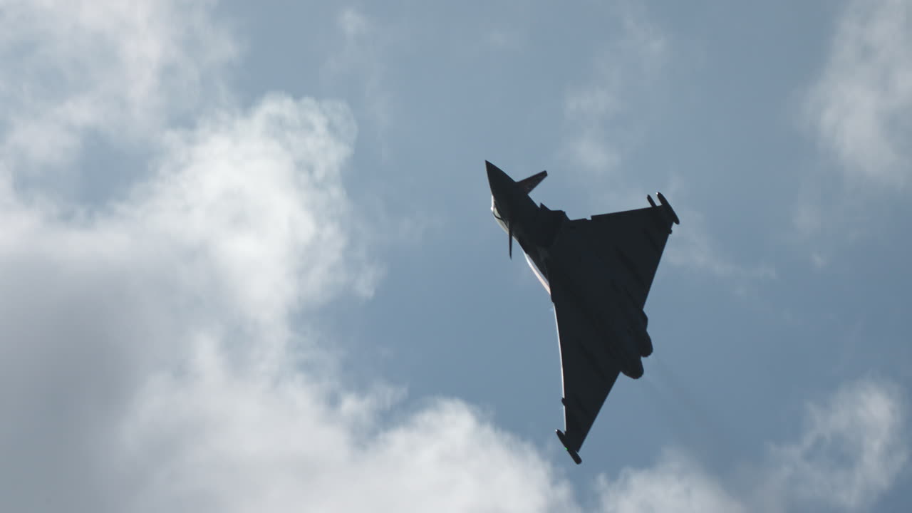 Silhouette of military jet fighter flying above in the sky with clouds in the background in slow motion