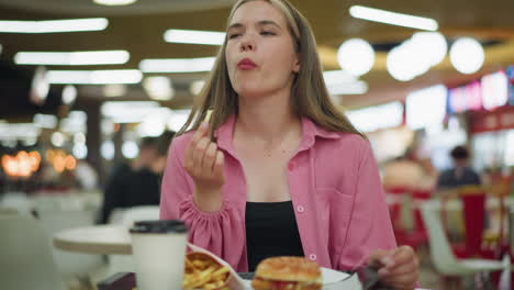 una señora sentada en un restaurante con café, papas fritas y hamburguesa en una bandeja delante de ella, toma un bocado de las papas fritas, cierra los ojos de satisfacción, y sacude la cabeza, disfrutando del delicioso sabor