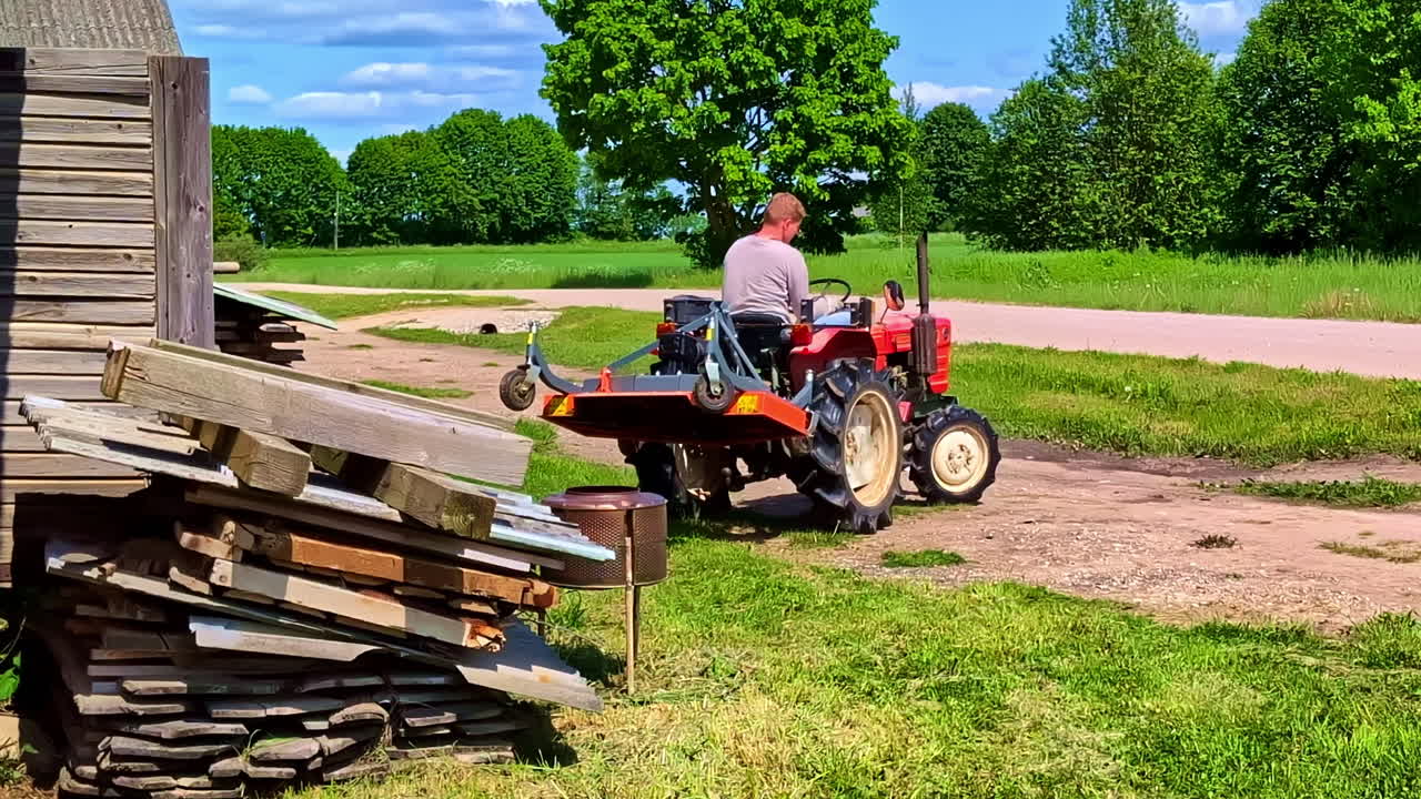 Man Riding Red Compact Tractor Past Stacked Timber Boards with Mower attached