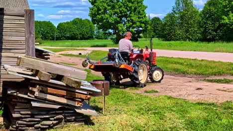 man riding red compact tractor past stacked timber boards with mower attached