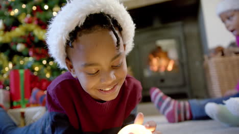 Feliz-Hijo-Afroamericano-Con-Sombrero-De-Navidad-Sosteniendo-Globo-De-Nieve,-Familia-En-Segundo-Plano,-Cámara-Lenta