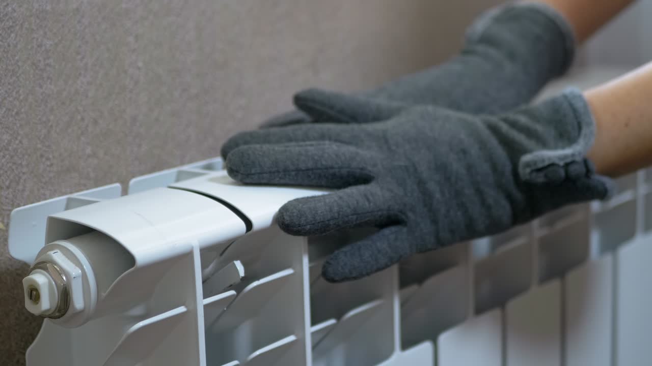 A Female in Woolen Gloves Warms Hands on a Radiator at Home in a Room. Zoom