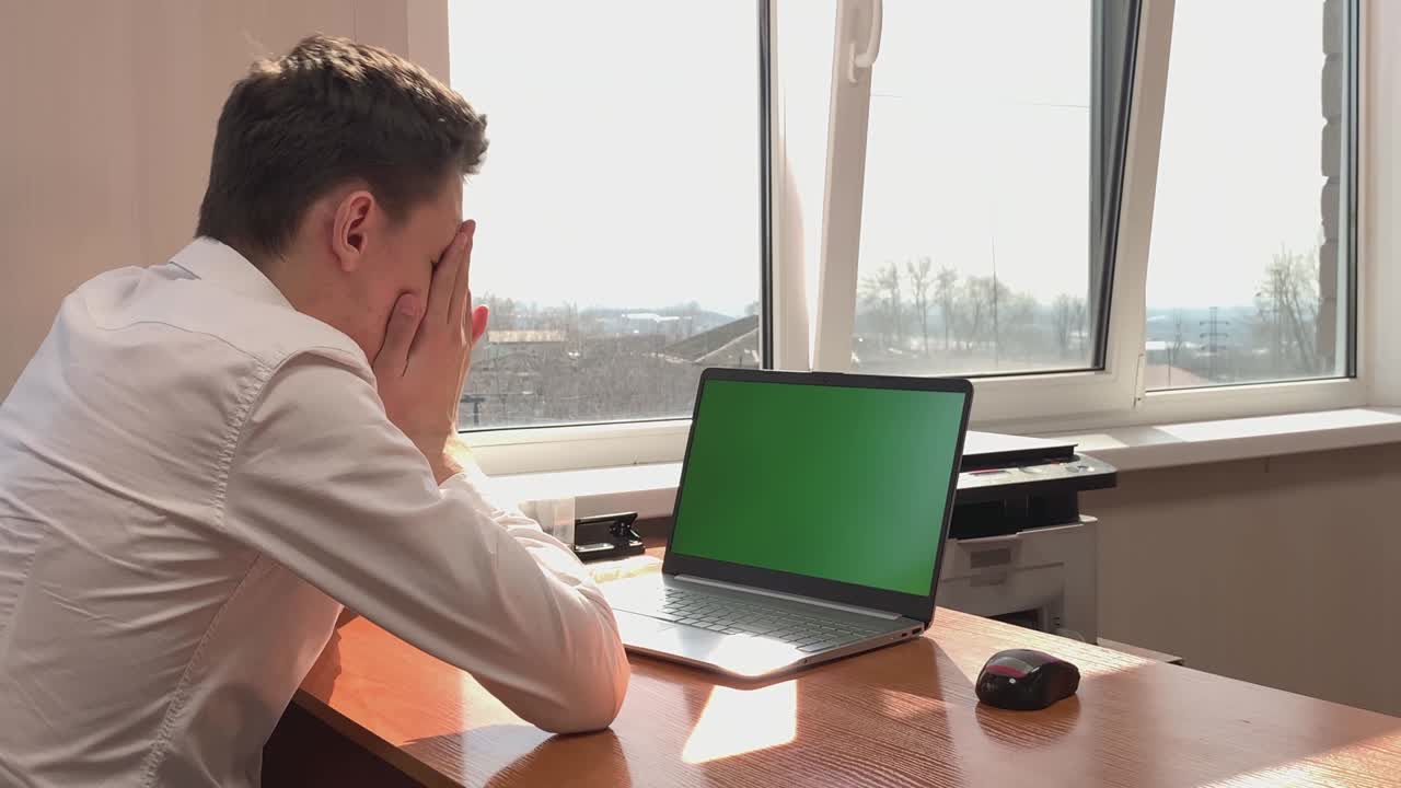 A young man enjoys success at work sitting at a desk in front of a computer with a green screen. Victory, success in work