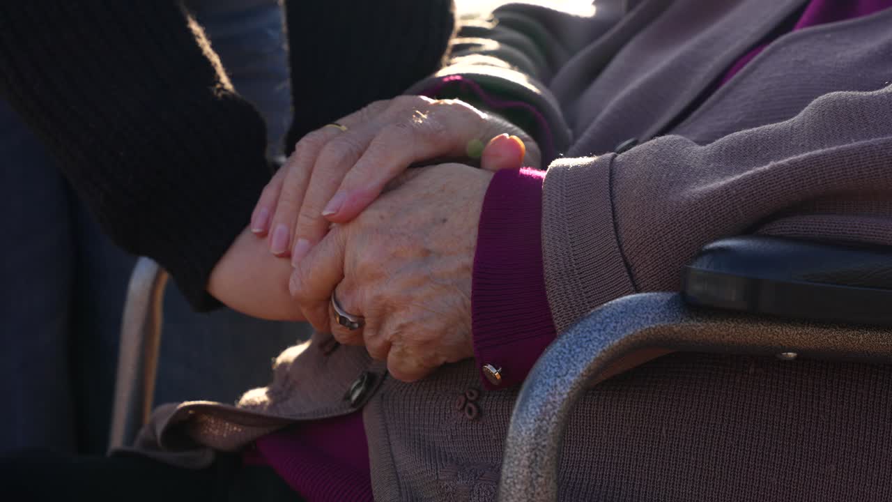 Granddaughter gently holding elderly woman’s hands in wheelchair. Close up